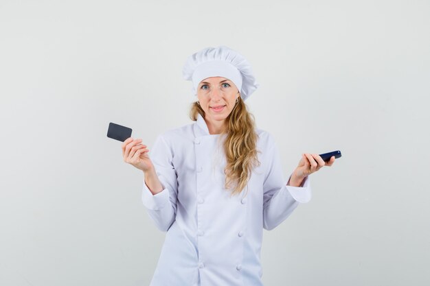 Femme chef tenant le téléphone portable et la carte en uniforme blanc et à la recherche optimiste.