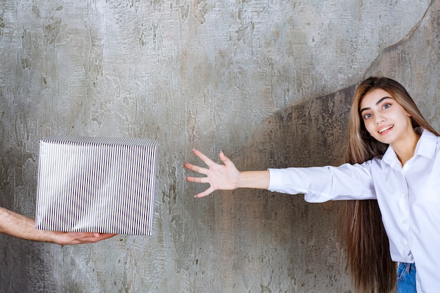 Une femme en chemise blanche debout sur un mur de béton se voit offrir une boîte-cadeau en argent et des mains impatientes de la prendre.