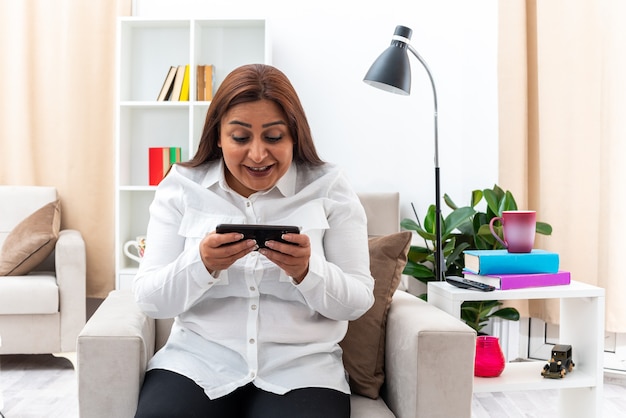 Femme en chemise blanche et pantalon noir jouant à des jeux à l'aide d'un smartphone heureux et excité assis sur la chaise dans un salon lumineux