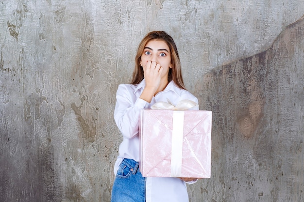 femme en chemise blanche tenant une boîte cadeau rose enveloppée de ruban blanc et a l'air effrayée ou terrifiée.