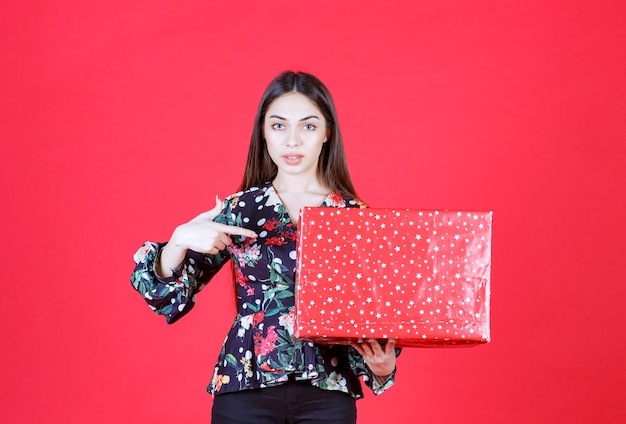 femme en chemise florale tenant une boîte-cadeau rouge avec des points blancs dessus.