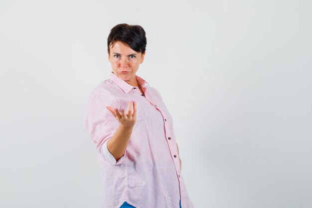 Femme en chemise rose qui s'étend de la main en geste perplexe, vue de face.