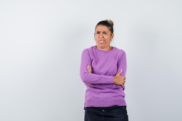 Femme en chemisier en laine debout avec les bras croisés, qui sort la langue et à la pensive