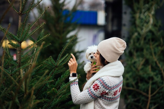 Femme avec un chien blanc dans ses bras près d'un arbre de Noël vert au marché