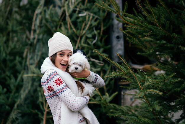 Femme avec un chien blanc dans ses bras près d'un arbre de Noël vert au marché