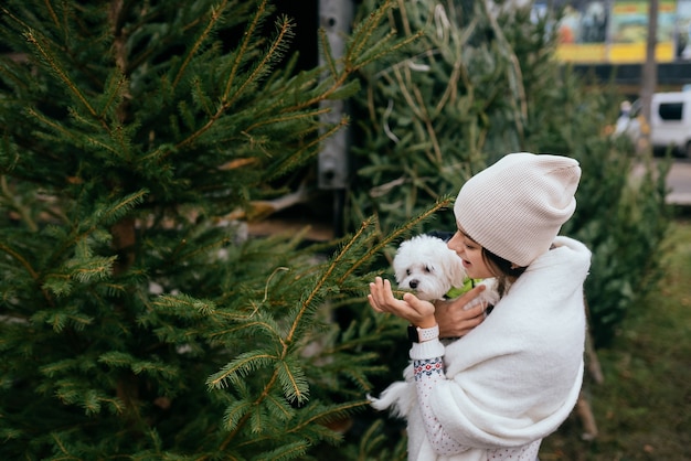 Femme avec un chien blanc dans ses bras près d'un arbre de Noël vert au marché