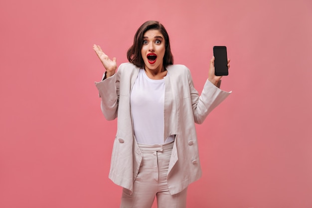 Femme choquée en costume de coton montre le téléphone sur fond rose. Femme surprise avec des lèvres brillantes dans des vêtements blancs tient le téléphone dans ses mains.