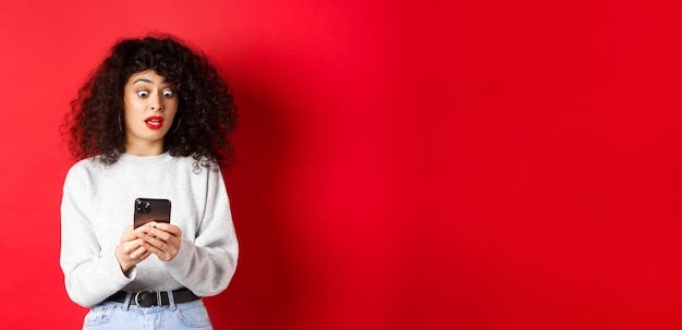 Photo gratuite une femme choquée regarde fixement l'écran du smartphone avec des yeux écarquillés lisant un message étrange debout sur une ba rouge