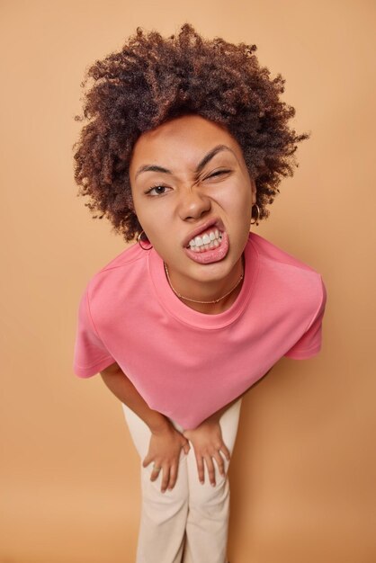 femme cligne des yeux serre les dents fait une grimace drôle à la caméra vêtue de vêtements décontractés se penche en avant isolé sur beige. Concept d'expressions de visage