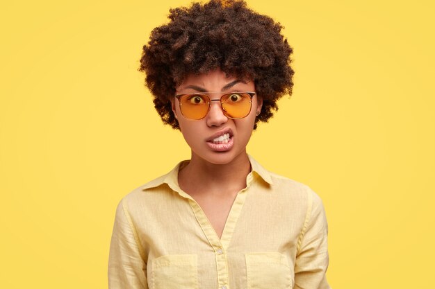 Femme en colère dans des lunettes de soleil serre les dents, regarde avec stress et expression irritée, soulève les sourcils, entend quelque chose de désagréable,