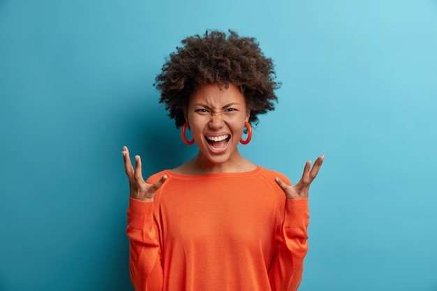 Femme en colère émotionnelle crie fort, exprime la haine et la rage, fait des gestes avec agacement, garde les mains levées de consternation, vêtue d'un pull orange, pose contre le motif bleu a une dispute avec son petit ami