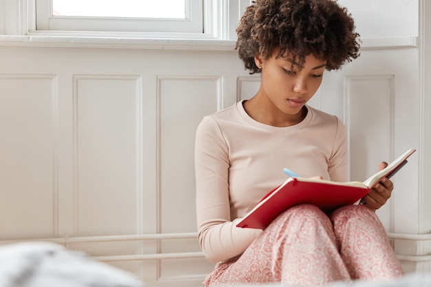 Photo gratuite femme concentrée écrit dans un cahier, utilise un stylo, vêtu de vêtements de nuit décontractés, est assis sur le sol à l'intérieur, prend des notes dans le journal,