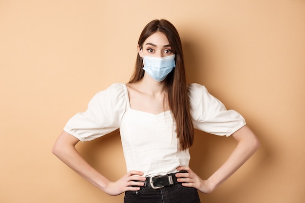 Femme de concept de covid et de style de vie dans un masque médical semblant alarmée devant la caméra debout sur un fond beige...