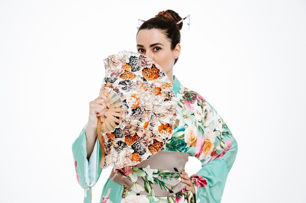 Femme confiante en kimono japonais traditionnel tenant la bouche d'un ventilateur à main avec elle sur blanc