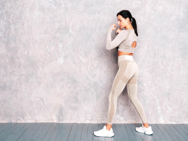 Femme confiante de remise en forme dans des vêtements de sport Sexy jeune beau modèle se tient dans un rack Femme posant près d'un mur gris en studio