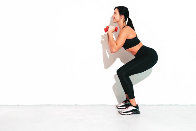 Femme confiante de remise en forme en vêtements de sport noirs Sexy jeune beau modèle avec un corps parfait Femme isolée sur un mur blanc en studio S'étirer avant l'entraînementFaire des squats avec des haltères