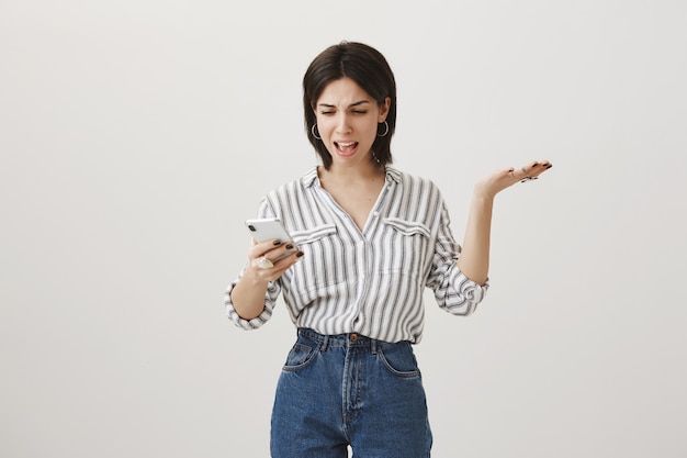 Femme confuse agacée regardant téléphone mobile et lever la main dans la consternation, se plaignant