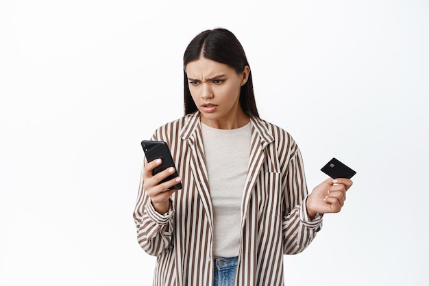 Femme confuse regardant son smartphone et tenant une carte de crédit en plastique ne peut pas comprendre où l'argent sur le compte bancaire vérifie le solde ou paie en ligne fond blanc