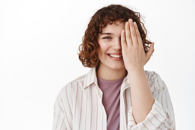 femme coning la moitié du visage avec la paume, souriante heureuse, vérifiant la vision dans le magasin d'un opticien ou montrant avant et après l'effet, debout sur blanc