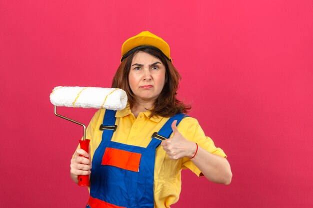 Femme constructeur mécontent portant des uniformes de construction et une casquette jaune tenant un rouleau à peinture en main montrant le pouce vers le haut sur un mur rose isolé