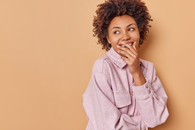 la femme contre la bouche avec la main sourit agréablement et détourne le regard se souvient de quelque chose de drôle porte une veste confortable concentrée derrière isolée sur un espace de copie vierge beige