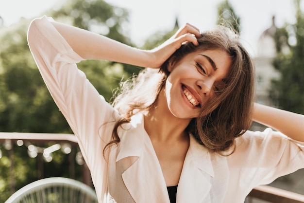 Femme cool en tenue légère riant sur la terrasse Fille brune bouclée en blouse beige souriant largement et posant sur le balcon