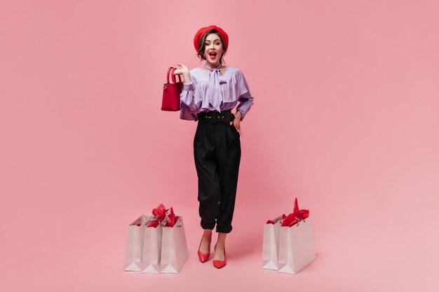 Femme coquette vêtue d'un pantalon élégant avec taille haute et béret lumineux pose avec petit sac à main et paquets sur fond rose.