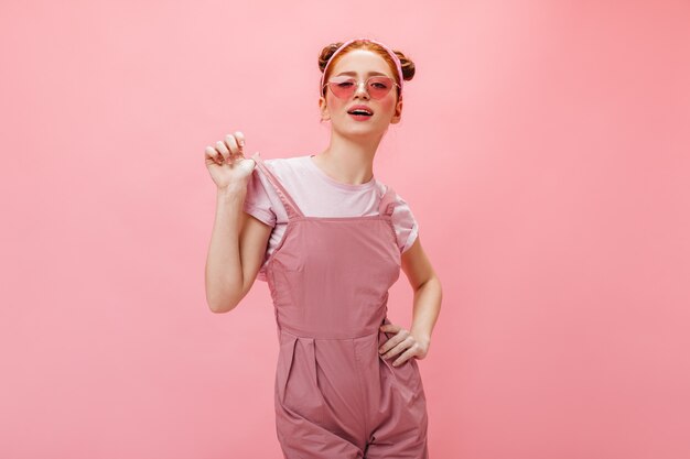 Femme coquine à lunettes et salopette se penche sur la caméra sur fond rose.