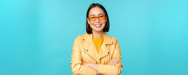 Femme coréenne élégante à lunettes de soleil et veste jaune bras croisés souriant et regardant confiant à la caméra debout sur fond bleu