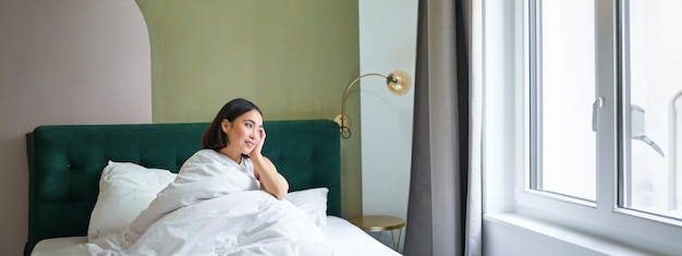 Une femme coréenne heureuse se réveille dans sa chambre d'hôtel, allongée dans son lit sous des couvertures chaudes et douillettes, des draps blancs