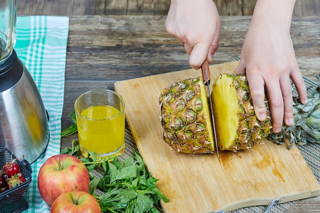 Femme coupant un ananas frais sur une planche à découper en bois à côté de pommes, verts et un verre de jus.
