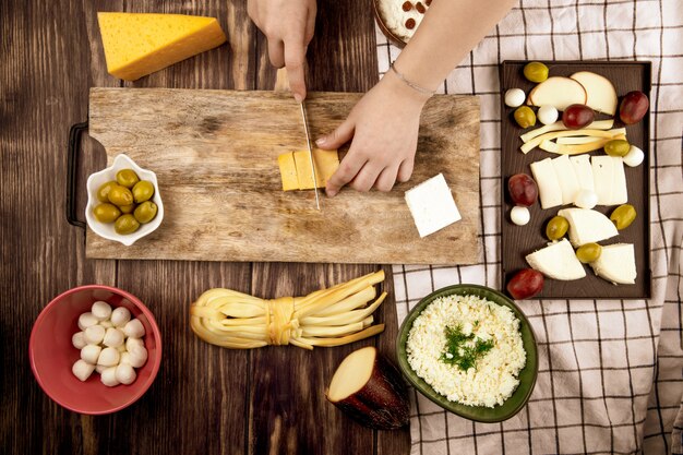 Une femme coupe le fromage hollandais sur une planche à découper en bois avec des olives marinées et divers types de fromage sur la vue de dessus rustique