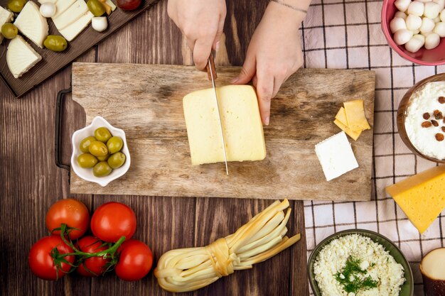 Une femme coupe le fromage hollandais sur une planche à découper en bois et des tomates fraîches olives marinées sur vue de dessus rustique