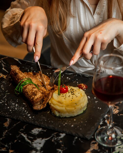 Femme coupe steak de viande avec pomme de terre masquée et verre de vin rouge.
