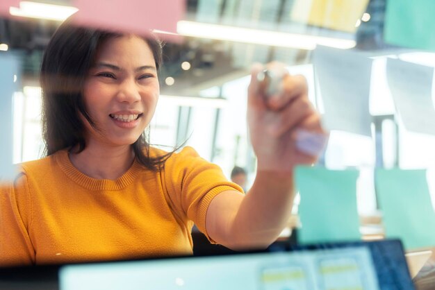 Une femme créative asiatique indépendante attrayante porte un point de main en tissu jaune décontracté et écrit un rappel de bâton de note papier dans l'espace de coworking nouveau style de vie avec une émotion réfléchie et fraîche