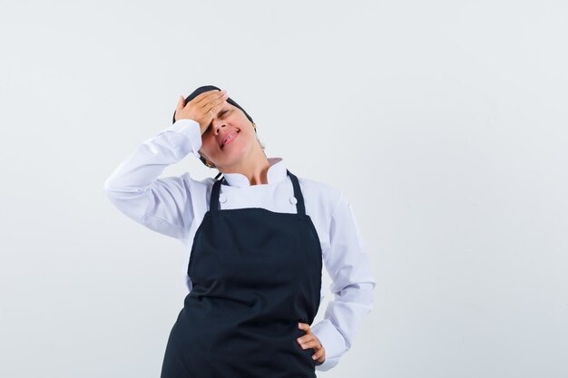 Femme cuisinier tenant la main sur le front en uniforme, tablier et élégant, vue de face.