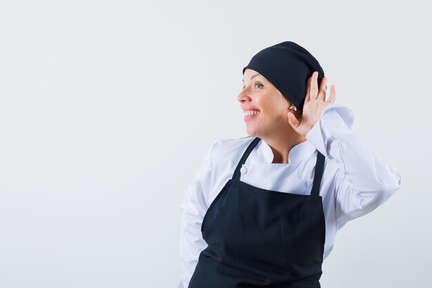 Femme cuisinier tenant la main près de l'oreille en uniforme, tablier et à la curieuse, vue de face.