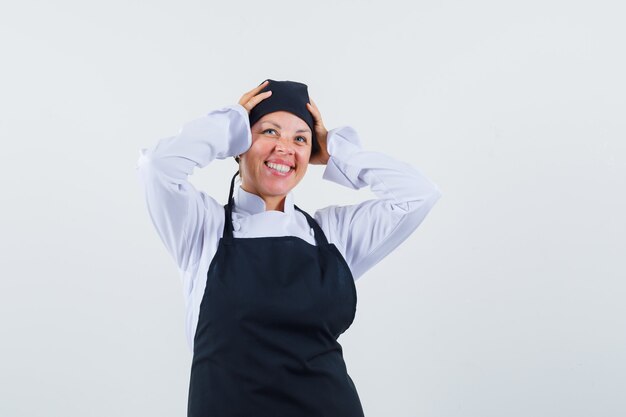 Femme cuisinier tenant les mains sur la tête en uniforme, tablier et à la joyeuse, vue de face.