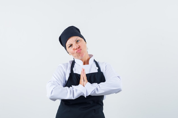 Femme cuisinière montrant le geste de namaste en uniforme, tablier et à la vue de face, hésitante.