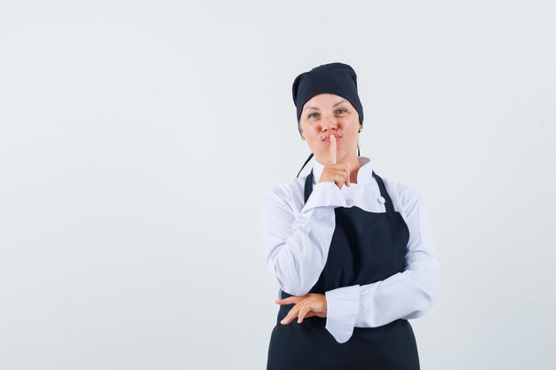 Femme cuisinière montrant le geste de silence en uniforme, tablier et à la recherche raisonnable. vue de face.