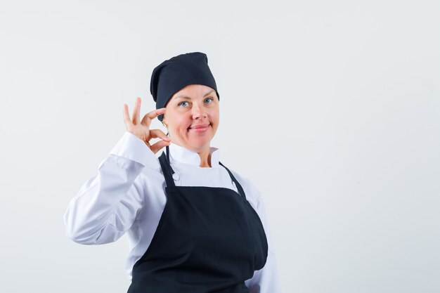 Femme cuisinière en uniforme, tablier montrant le geste correct et à la vue confiante, vue de face.