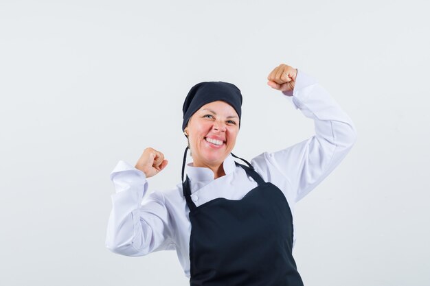 Femme cuisinière en uniforme, tablier montrant le geste du gagnant et à la béatitude, vue de face.