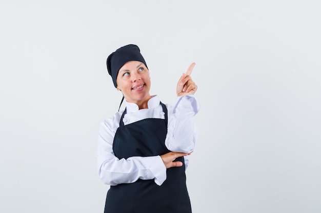 Femme cuisinière en uniforme, tablier pointant vers le haut et à la rêveuse, vue de face.