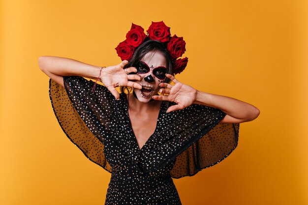 Une femme dangereuse avec un masque de crâne essaie de faire peur. Photo de fille avec des roses dans ses cheveux sur fond orange.