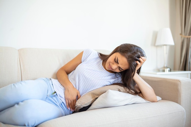 Femme dans l'expression douloureuse tenant la main contre le ventre souffrant de période menstruelle painsitting triste sur le lit à la maison ayant une crampe abdominale dans le concept de santé féminine