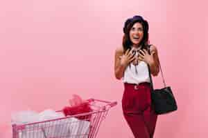 Photo gratuite femme dans une joyeuse surprise regarde la caméra et pose à côté du chariot rose. dame en chemisier blanc et pantalon lumineux rit sur fond isolé.