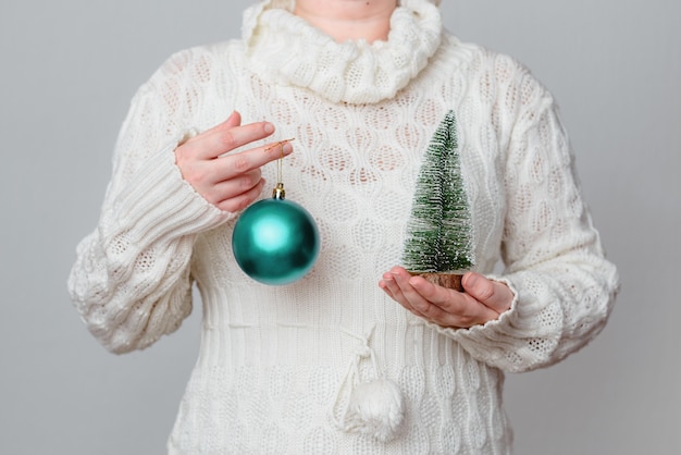 Femme dans un pull blanc tenant une boule de Noël turquoise et pin miniature