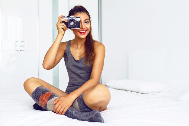 Femme dans des vêtements décontractés confortables, assis sur un grand lit blanc et prendre des photos sur un appareil photo vintage