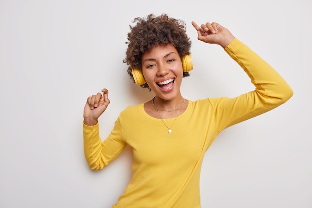 la femme danse avec les bras levés porte un pull jaune décontracté et des écouteurs sans fil ont une humeur optimiste isolée sur blanc. Émotions des gens et concept de passe-temps