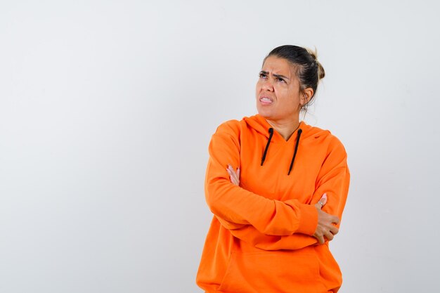 Femme debout avec les bras croisés en sweat à capuche orange et à la sombre, vue de face.